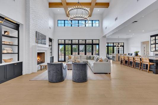 Open-concept luxury custom home living room with double-height ceilings, exposed wood beams, iron ring chandelier, white brick fireplace, grey upholstered seating, and black-framed windows overlooking a dining area