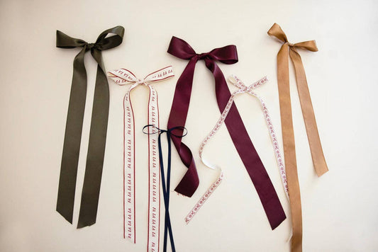 Flat lay of elegant satin hair bows in olive green, deep burgundy, and camel with printed ribbon accents on a neutral background
