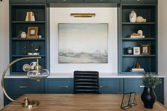 Styled built-in shelving with dark teal cabinetry, curated home decor accessories, framed art with brass picture light, and a black leather chair in a designer home office