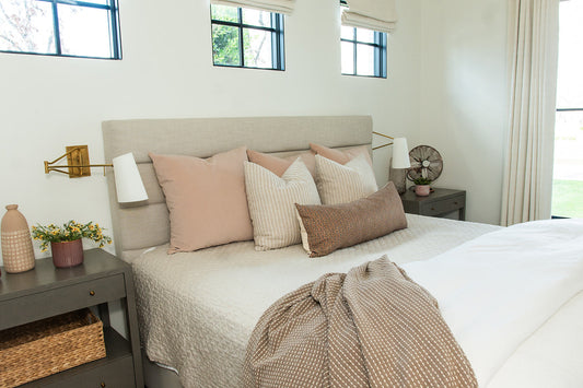 Beautifully styled bedroom with upholstered linen headboard, layered neutral throw pillows, brass wall sconces, sage green nightstands, and a vintage fan on the bedside table