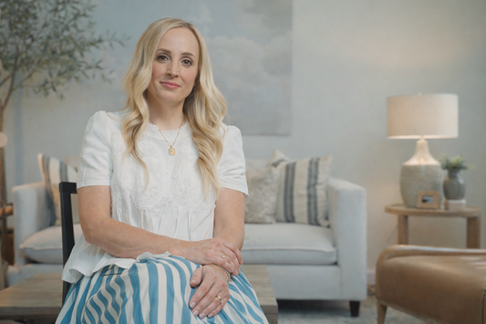 Ashley Kuhni, founder and interior designer of Designly Done, sitting in a styled living room with soft neutral decor and a table lamp in the background