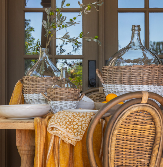 Close-up of curated home decor vignette with clear glass demijohn vases, olive branch greenery, and a woven rattan basket near a window with natural light