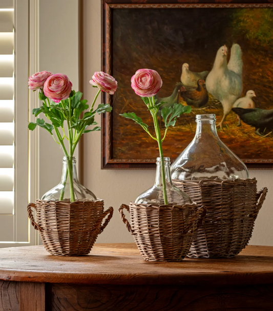 Boutique home decor vignette with pink ranunculus flowers in a glass vase, a vintage oil painting of white hens in a dark wood frame, and a clear glass bottle on a neutral backdrop