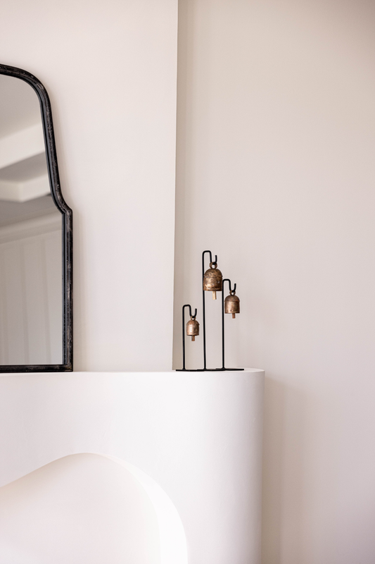 Minimalist home decor vignette with an arched black metal mirror, vintage brass bell on an iron stand, and warm neutral wall in a boutique setting