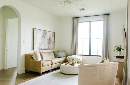 Bright living room corner with beige sofa, abstract landscape wall art, sheer floor-length curtains, and a styled coffee table with decorative accents